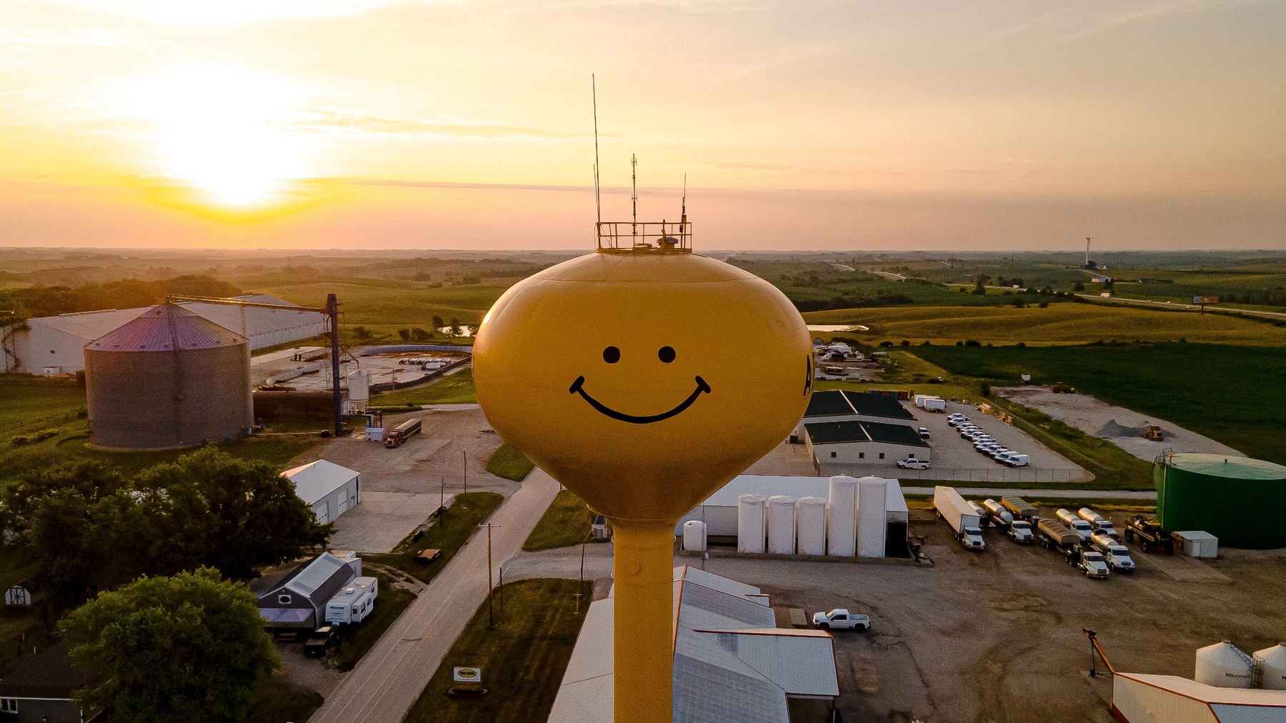 Aerial drone scenery photo of a yellow smiley face water tower at sunrise over western Iowa fields and buildings
